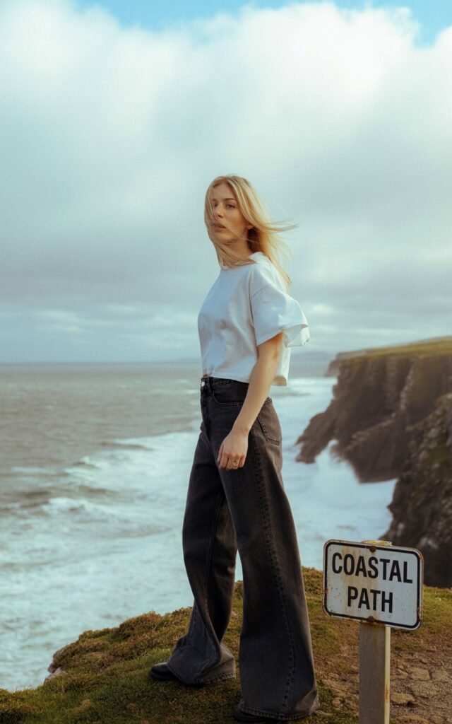 A blonde model stands beneath a misty sky on the windswept cliffs of Ireland, her oversized white tee loosely tucked into dark, structured wide-leg jeans. Wind ruffles her hair as waves crash dramatically below. The mood is cinematic and powerful, with a sense of quiet freedom.