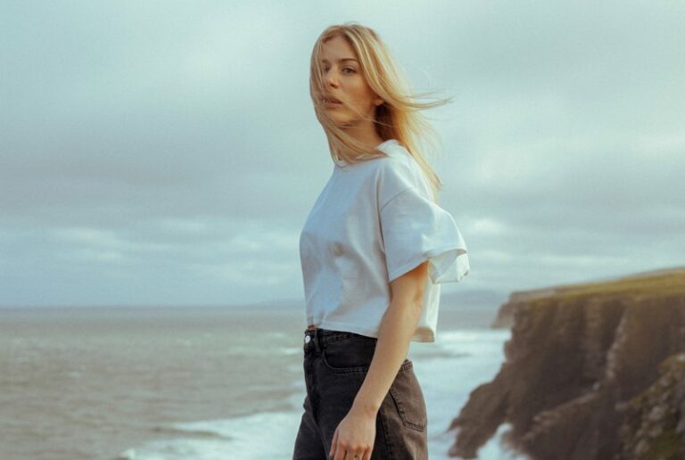 A blonde model stands beneath a misty sky on the windswept cliffs of Ireland, her oversized white tee loosely tucked into dark, structured wide-leg jeans. Wind ruffles her hair as waves crash dramatically below. The mood is cinematic and powerful, with a sense of quiet freedom.