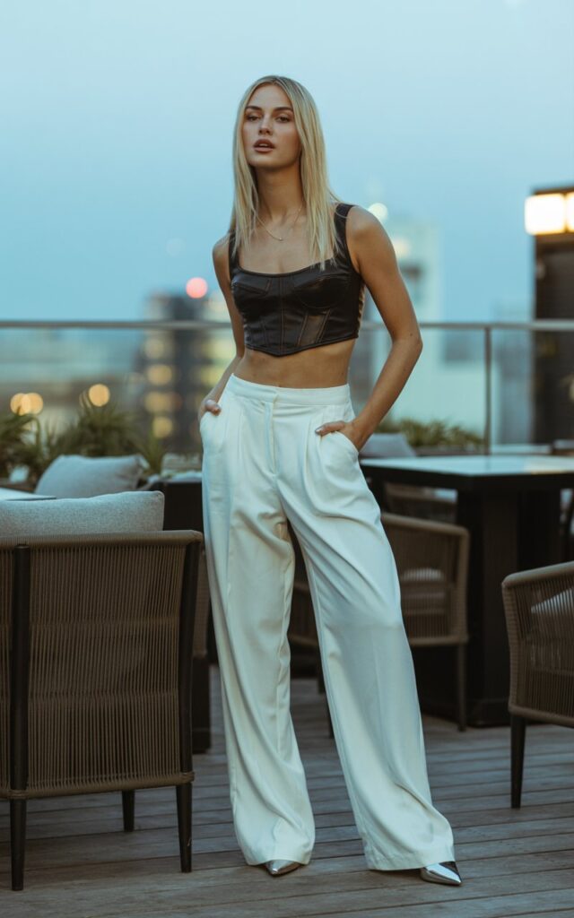 A blonde model with sleek straight hair and sharp cheekbones poses under soft daylight in a chic rooftop bar. She wears a black cropped corset top with high-waisted white wide-leg trousers, paired with pointed-toe pumps. Standing with one hand in pocket, gazing directly at the camera with boss energy.
