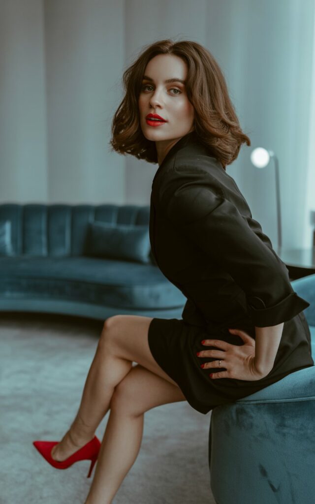 A brunette with shoulder-length waves poses in a chic indoor lounge under soft spotlighting. She wears a timeless black mini dress styled with bold red stilettos and a matching red lip. Standing with one hand on her hip, giving a sultry yet classy gaze.