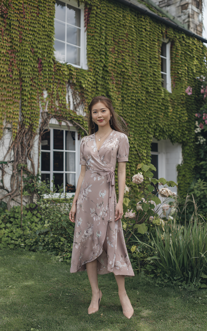 A light-skinned East Asian woman standing in front of an ivy-covered English cottage, wearing a muted floral wrap dress with nude pumps and delicate gold jewelry.