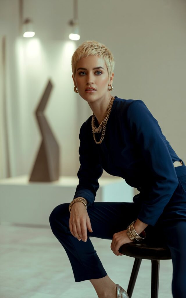 A short-haired blonde with sharp features poses in a modern indoor studio with warm spotlighting. She wears a fitted navy jumpsuit styled with chunky layered gold chains, bangles, and metallic heels. Sitting on a stool, leaning slightly forward with an elegant, powerful gaze.