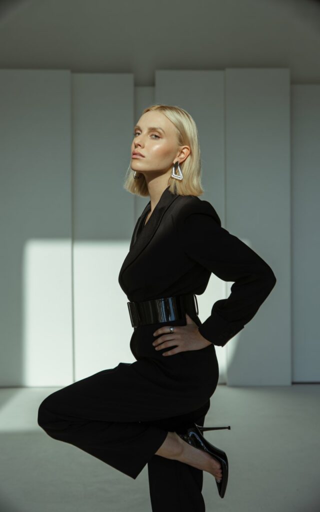 A tall blonde model stands in a modern studio with geometric props. She wears a black fitted jumpsuit cinched with a wide leather belt, stilettos, and bold earrings. Dramatic side lighting highlights her sharp jawline. She poses with one hand on her waist, chin lifted, radiating confidence.