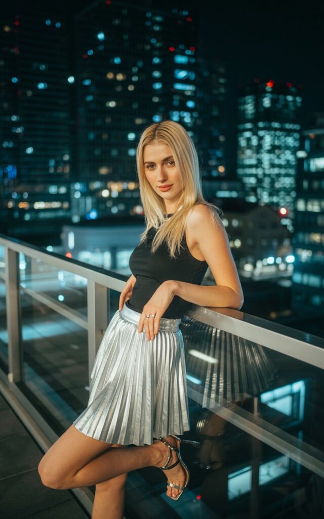 A tall blonde with sleek straight hair poses at an evening rooftop setting with city lights. She wears a silver metallic pleated skirt paired with a fitted black tank and strappy heels. Leaning casually against a railing, natural smile, outfit catching the glow of city lights.