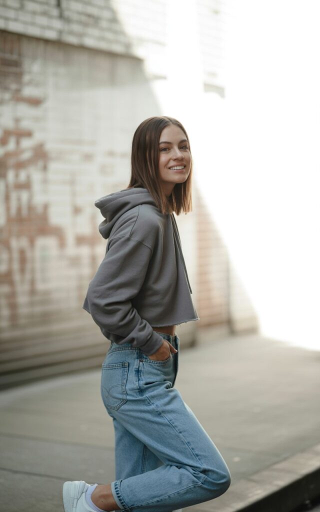 A white-skinned female model with sleek straight brown hair, wearing a cropped grey hoodie and light blue high-waisted jeans with white sneakers. Shot on an urban street with brick walls in the background, natural daylight. She stands casually with one hand in her pocket, smiling effortlessly, giving off a chill, put-together vibe.