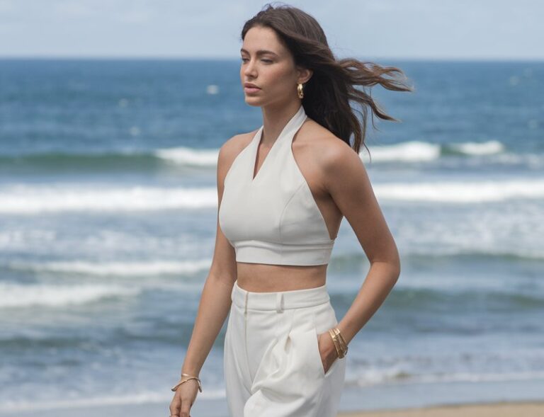 Beach promenade setting with ocean in the background. The model wears white wide-leg trousers with a halter crop top, flat sandals, and gold jewelry. She walks barefoot on the boardwalk, hair flowing, carefree expression.