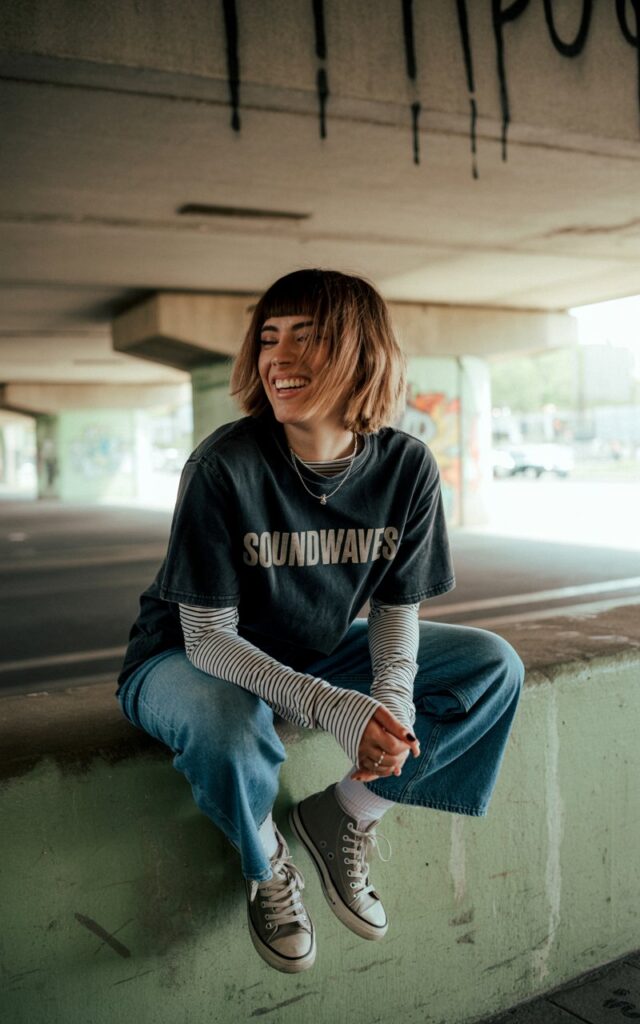 City underpass setting. Brunette with layered shag haircut wears a striped long-sleeve under a black band tee, distressed jeans, and high-top sneakers. She leans against a concrete wall, one foot propped behind her, laughing naturally.