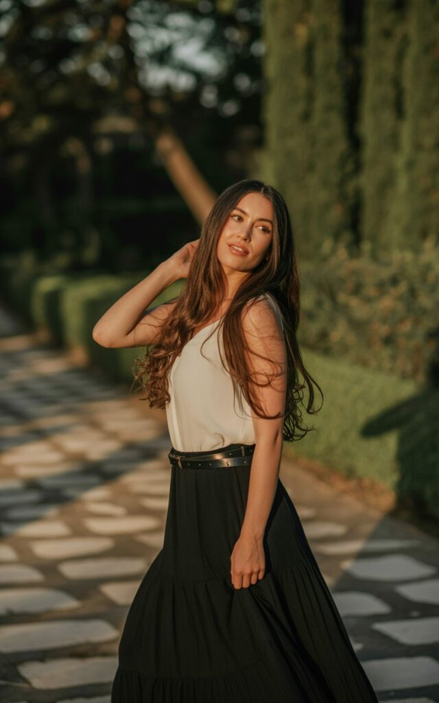 Full-body of a tall brunette in a flowing black maxi skirt and fitted white blouse, cinched with a slim belt. Shot at golden hour in a garden path. Model standing gracefully, hair cascading, serene expression.