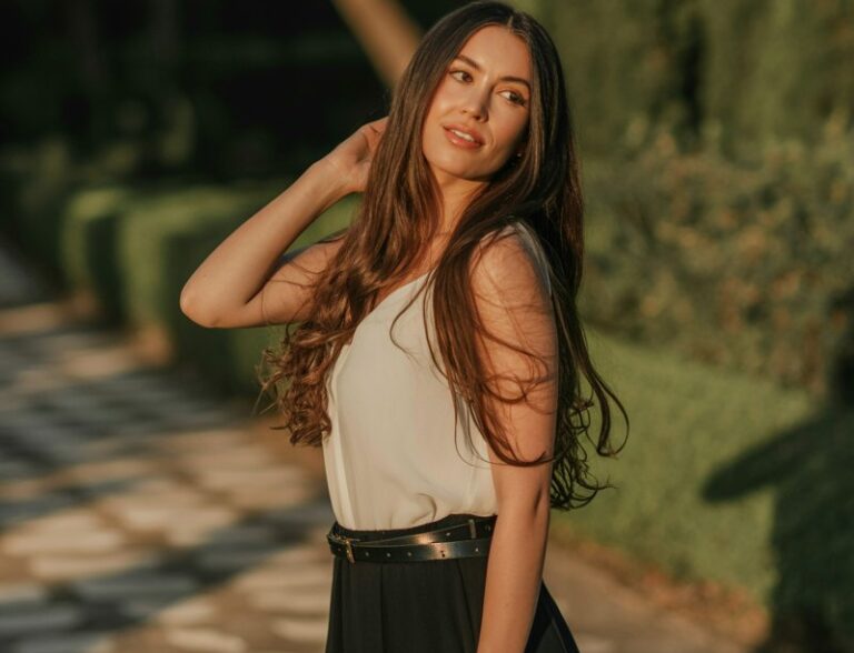 Full-body of a tall brunette in a flowing black maxi skirt and fitted white blouse, cinched with a slim belt. Shot at golden hour in a garden path. Model standing gracefully, hair cascading, serene expression.