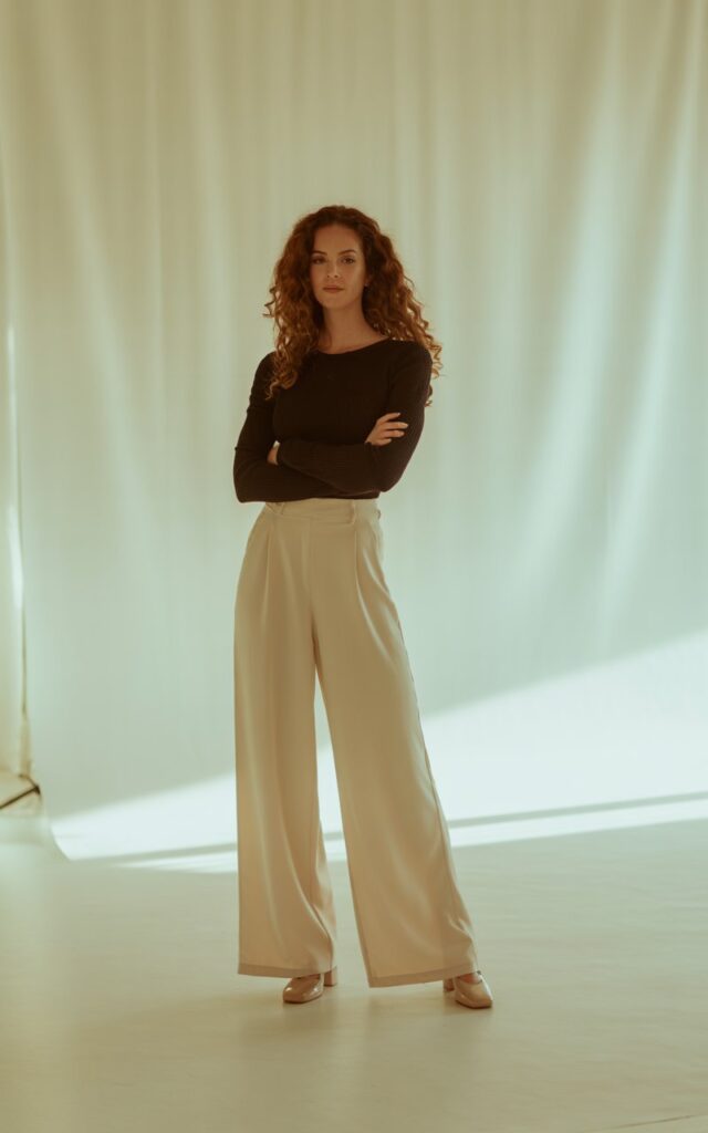 Full-body of a tall brunette in wide cream palazzo pants with a fitted black knit top, chunky heels. Shot indoors with warm studio light. Hair styled naturally voluminous. Model standing tall, arms crossed lightly, strong expression.
