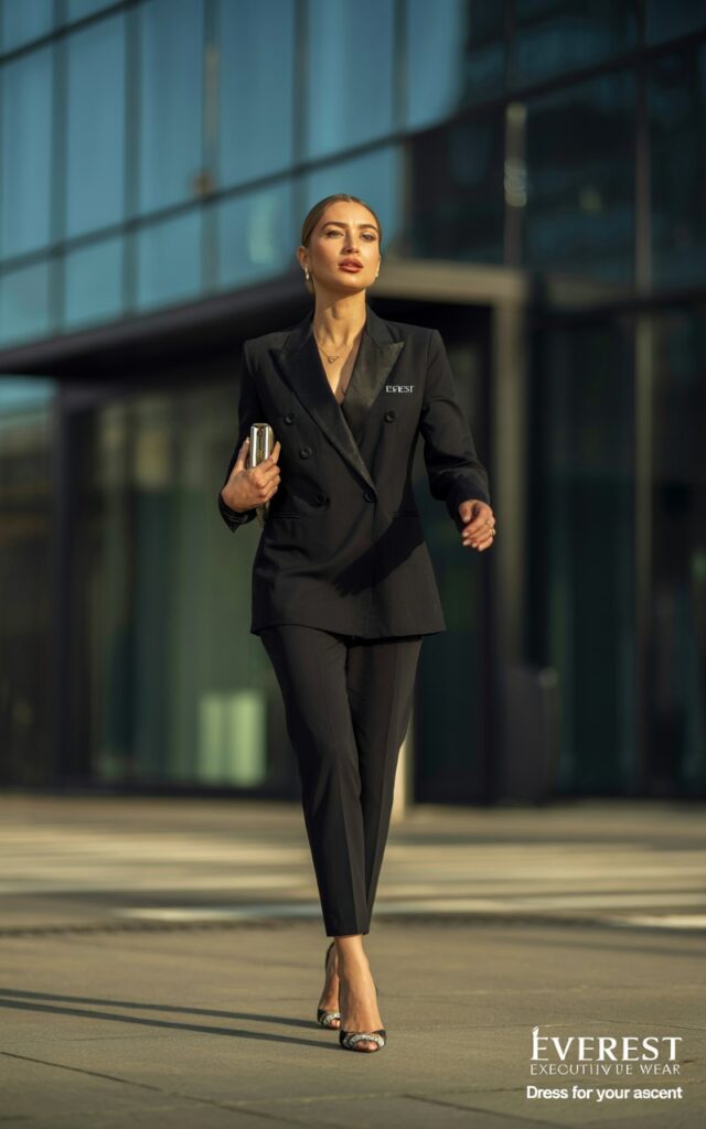Full-body shot in a sleek corporate building entrance with natural daylight. The model wears a black double-breasted blazer with slim cigarette pants, stilettos, and a small clutch. Hair straightened, makeup bold but elegant. She walks forward with a powerful stride, exuding authority.