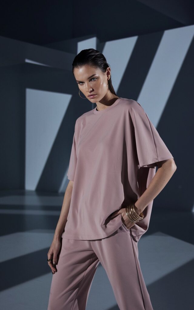 In a futuristic, all-white studio with sharp shadows and geometric backdrops, a statuesque model wears a dusty pink monochrome loungewear set. Subtle gold hoops and layered bracelets shimmer in the spotlight. The aesthetic is minimal, high-fashion, and editorially clean.