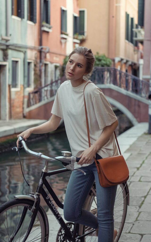 In a pastel-toned Venice canal alleyway, a soft-featured model leans on a bicycle, wearing a tucked-in oversized white tee and faded skinny jeans. Her leather crossbody hangs at her hip. Warm afternoon light reflects off the water. The atmosphere is relaxed, feminine, and charmingly cinematic.
