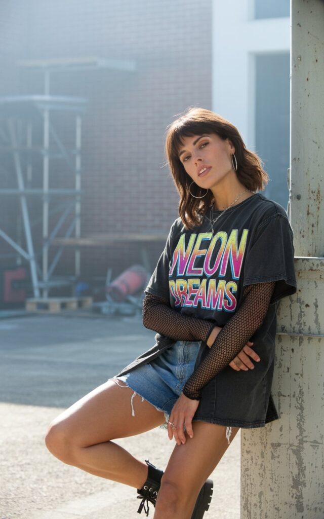 Industrial warehouse setting with moody daylight streaming in. Brunette model with bangs wears a black mesh long-sleeve under a baggy oversized graphic tee, paired with distressed shorts and chunky boots. She leans against a concrete pillar, arms crossed, expression rebellious yet playful.