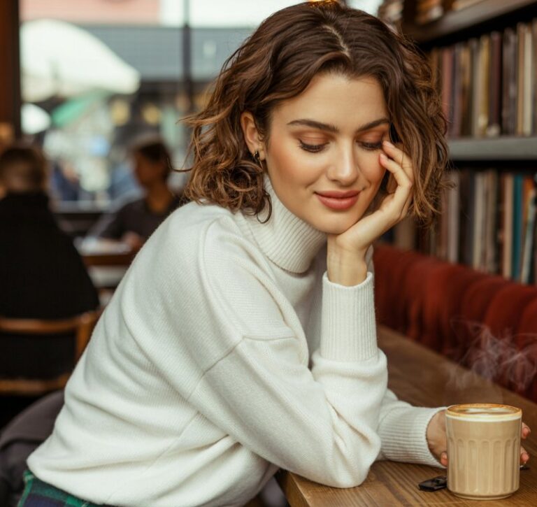 Lifestyle capture of a brunette with natural waves, wearing a classic plaid mini skirt, cream turtleneck, and ankle boots. Setting cozy café interior with warm window light streaming through. She sits at a wooden table with coffee, chin resting in one hand, giving a soft candid smile.