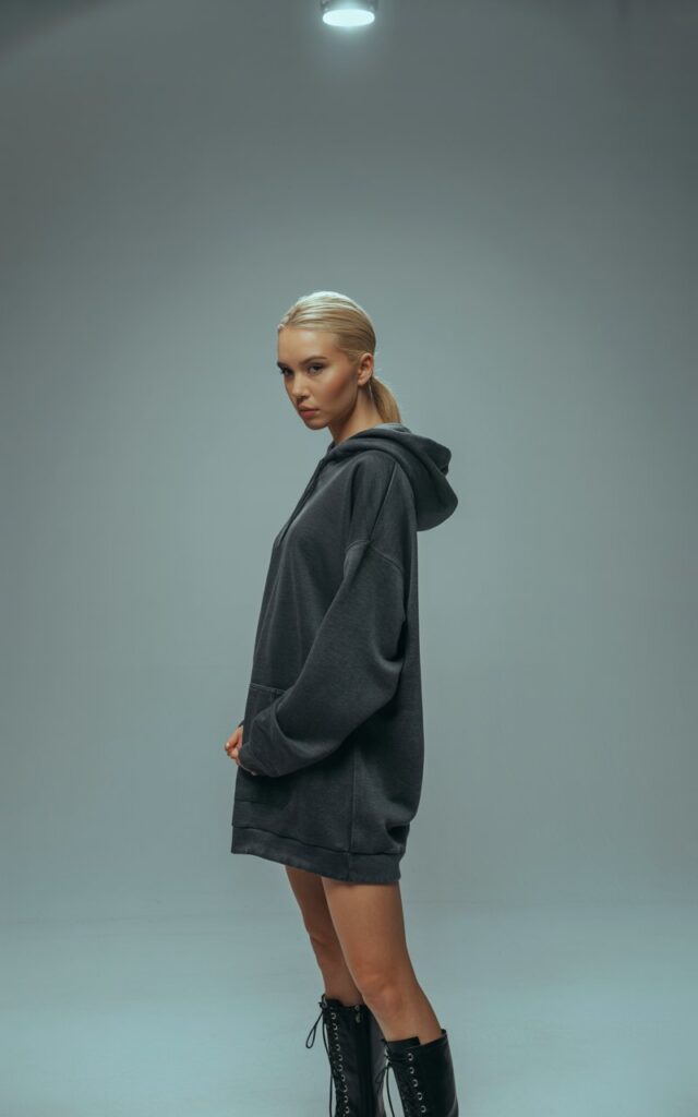 Minimal studio backdrop with soft natural light. Blonde model wears a charcoal oversized hoodie dress with black knee-high lace-up boots. Hair in a sleek high ponytail. She stands tall with one leg slightly forward, expression strong and confident.