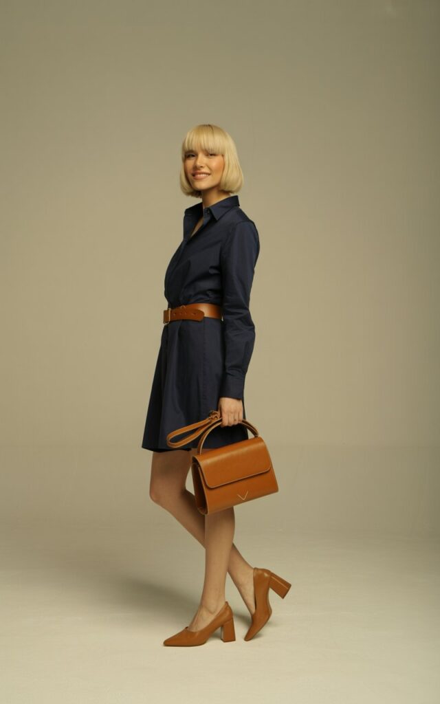 Model with blonde bob haircut, shot in studio with neutral backdrop and soft lighting. She wears a navy shirt dress cinched at the waist with a tan belt, paired with tan block heels and a structured leather handbag. Standing tall, one leg slightly bent, looking playful.