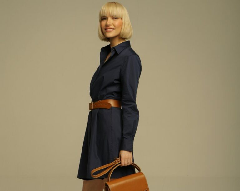 Model with blonde bob haircut, shot in studio with neutral backdrop and soft lighting. She wears a navy shirt dress cinched at the waist with a tan belt, paired with tan block heels and a structured leather handbag. Standing tall, one leg slightly bent, looking playful.