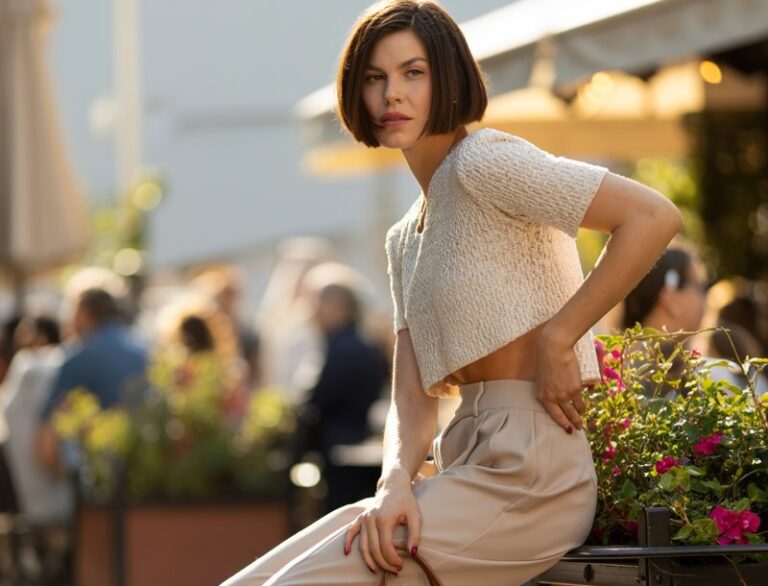 Model with sharp bob haircut, posing on an outdoor café terrace at golden hour. She wears high-waisted beige wide-leg trousers paired with a cropped white knit sweater, finished with chunky sneakers. A leather tote sits beside her. Standing confidently with one hand on her hip, chin slightly tilted, editorial vibe.