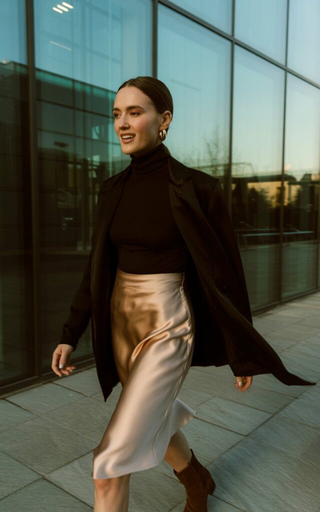 Model with sleek brunette hair parted in the middle, shot against modern glass architecture at sunset. She wears a champagne slip skirt with a black fitted turtleneck tucked in, ankle boots, and minimal jewelry. Walking mid-step, candid smile, natural vibe.