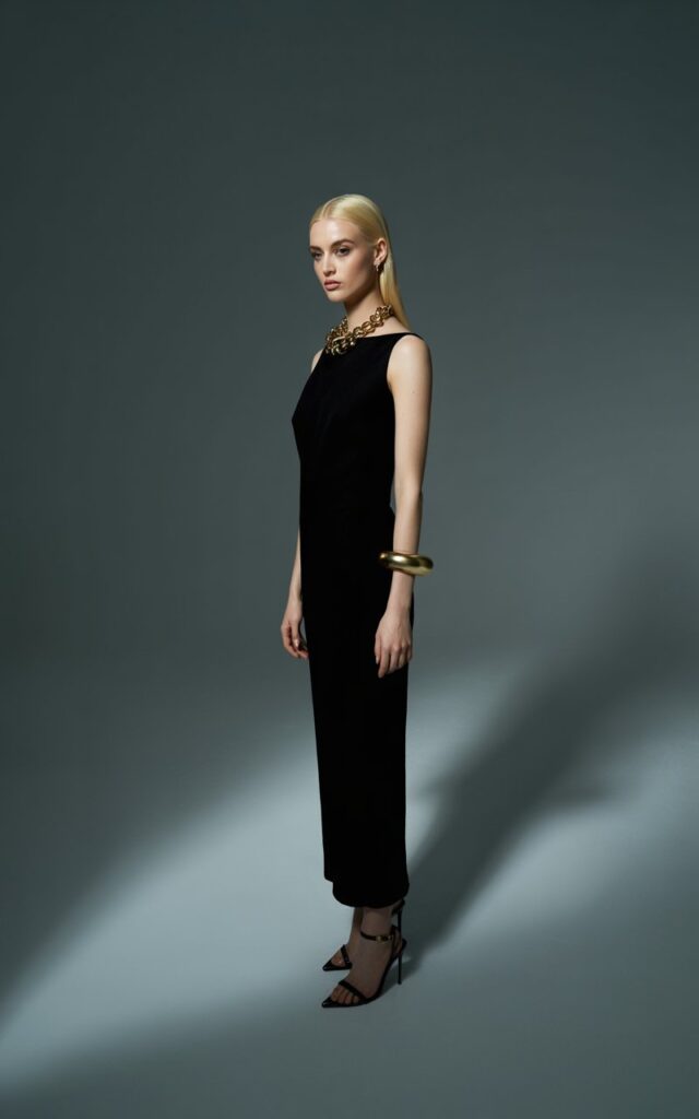 Model with sleek long blonde hair parted center. Outfit fitted black column dress, bold chunky gold necklace, and stiletto heels. Setting elegant indoor studio with dramatic spotlighting. Pose standing tall, head slightly tilted, expression elegant and poised.