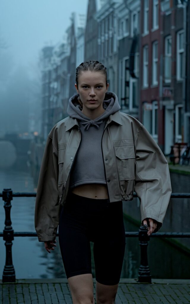 Set in the foggy streets of an early morning Amsterdam canal, a brunette model with chiseled features walks briskly in a sleek taupe utility jacket layered over a cropped hoodie and black biker shorts. The light is cool and moody. The look is urban, athletic, and ultra-modern.