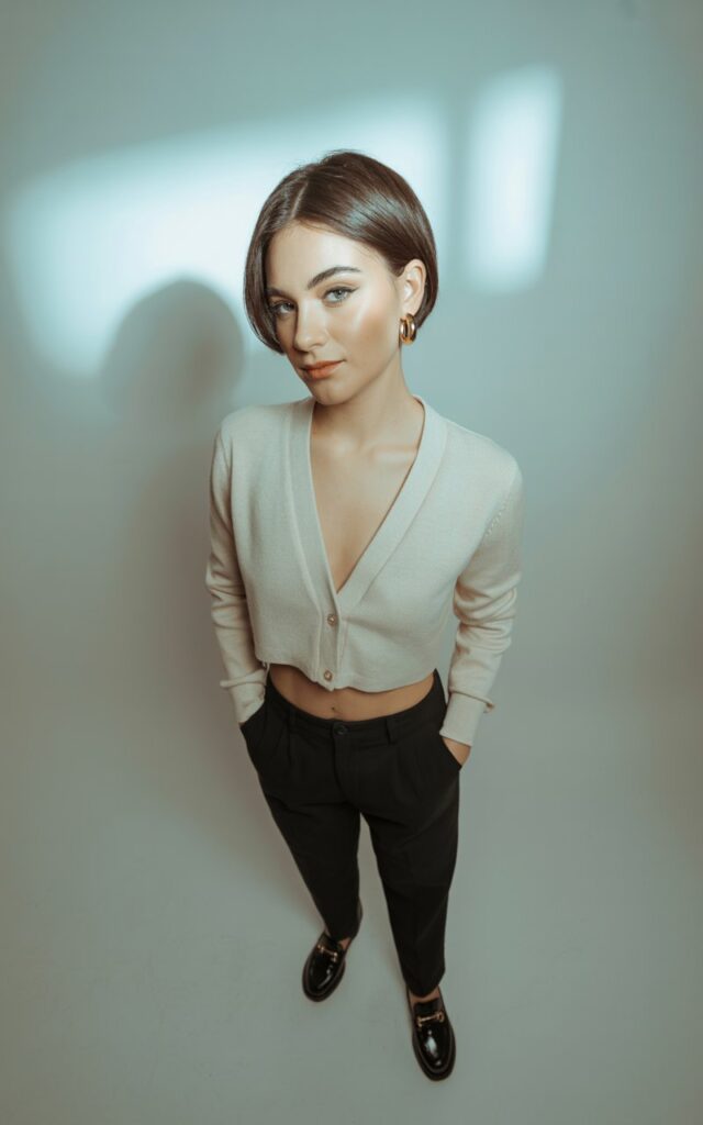 Studio background with soft grey backdrop and controlled lighting. Model wears a cropped cream cardigan buttoned halfway, paired with slim tailored black trousers and black loafers. Minimal gold jewelry. Hair is sleek in a short bob. Pose standing with hands in pockets, looking poised and sharp.
