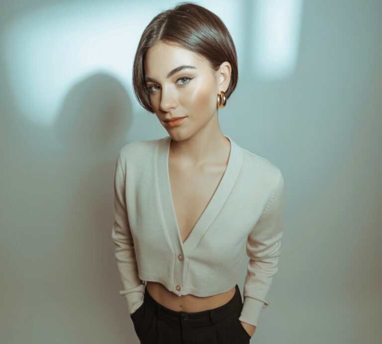 Studio background with soft grey backdrop and controlled lighting. Model wears a cropped cream cardigan buttoned halfway, paired with slim tailored black trousers and black loafers. Minimal gold jewelry. Hair is sleek in a short bob. Pose standing with hands in pockets, looking poised and sharp.