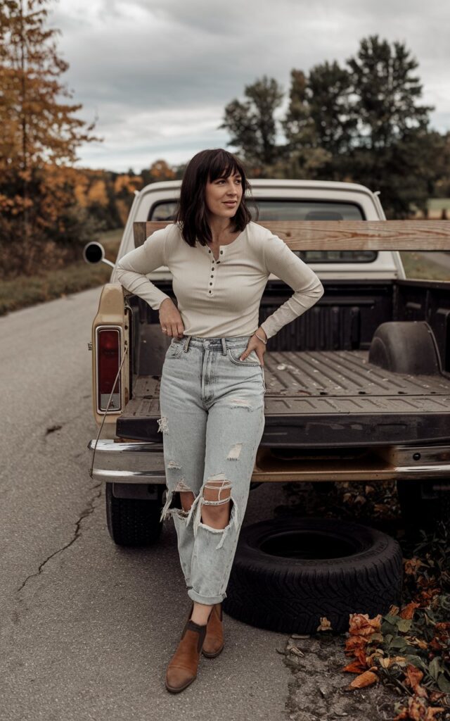 White-skinned European woman leaning against a vintage pickup truck, wearing a cream Henley shirt, distressed boyfriend jeans, brown ankle boots, rural road with autumn trees behind.
