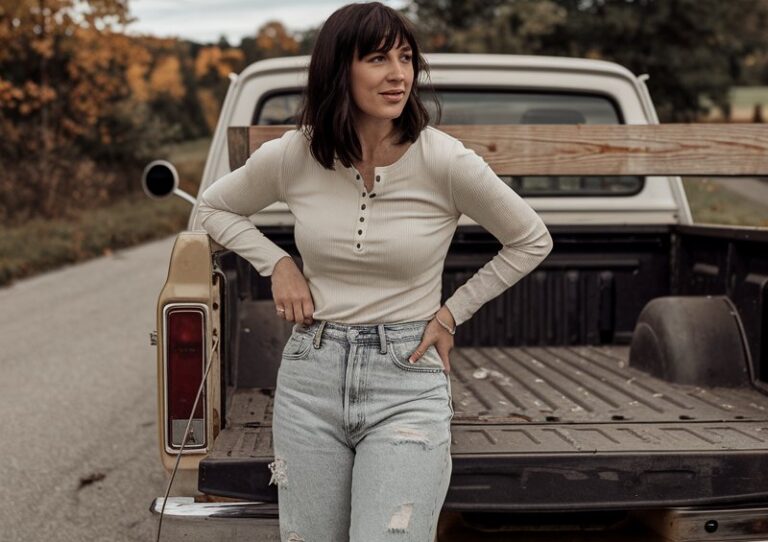 White-skinned European woman leaning against a vintage pickup truck, wearing a cream Henley shirt, distressed boyfriend jeans, brown ankle boots, rural road with autumn trees behind.