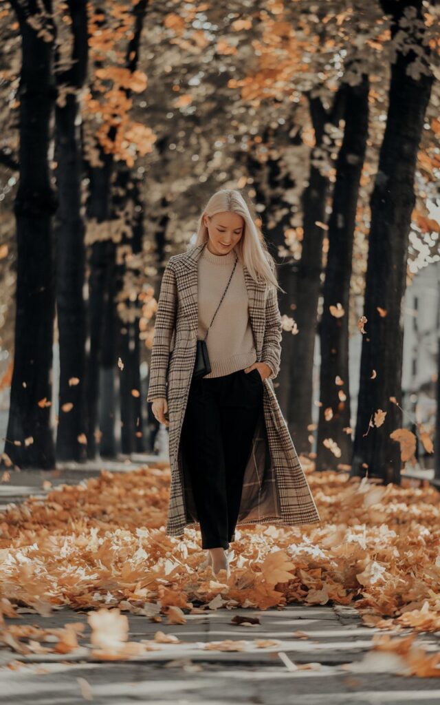 White-skinned European woman strolling down a tree-lined avenue, beige sweater and black pants under a long plaid coat, crossbody bag, golden autumn leaves swirling in the wind, warm afternoon sunlight.