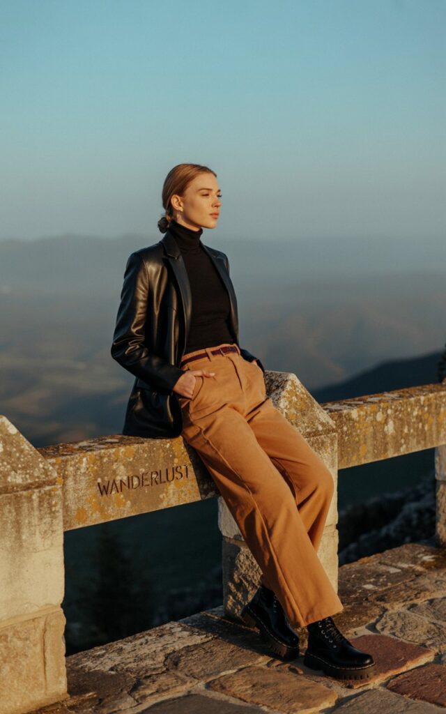 A fair-skinned model leans against a mountain railing at golden hour. She wears a black turtleneck tucked into wide-leg camel pants, paired with chunky black boots. She rests one foot on the railing, hands in pockets, gazing into the distance with a calm expression. The warm sunlight highlights the outfit’s textures and natural details.