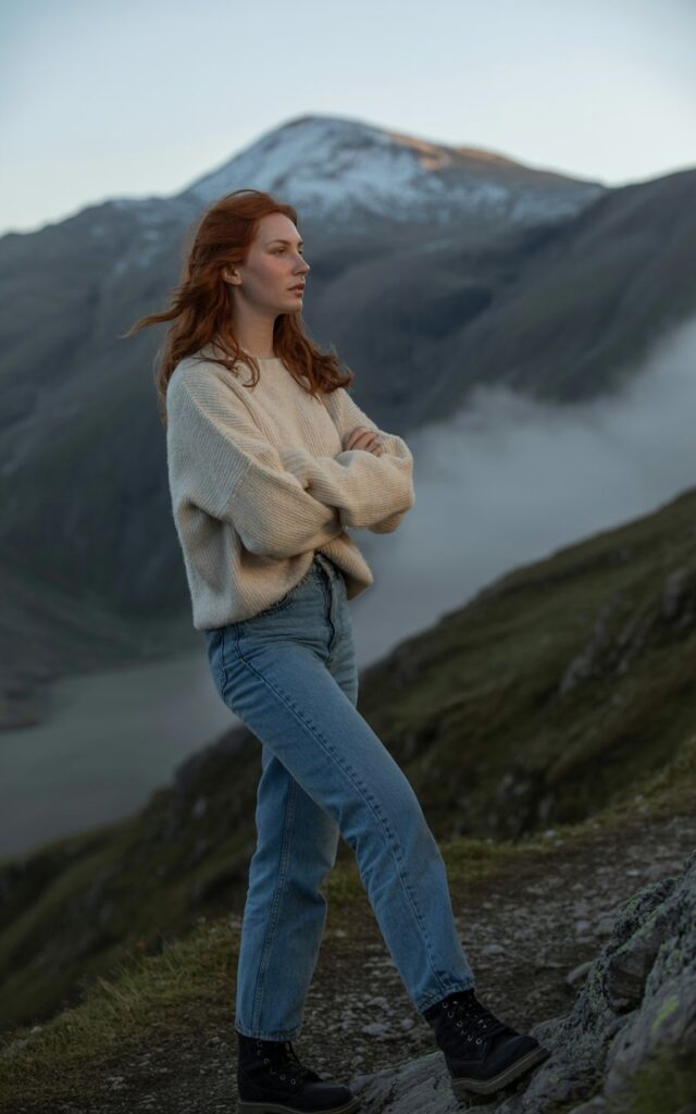 A fair-skinned model stands on a rugged mountain path at soft afternoon light. She wears a cozy cream knit sweater, high-rise blue jeans, and black combat boots. She stands with arms crossed, gazing thoughtfully into the distance. The soft light catches the knit’s texture and her wind-blown hair, while the mountain landscape frames the shot.