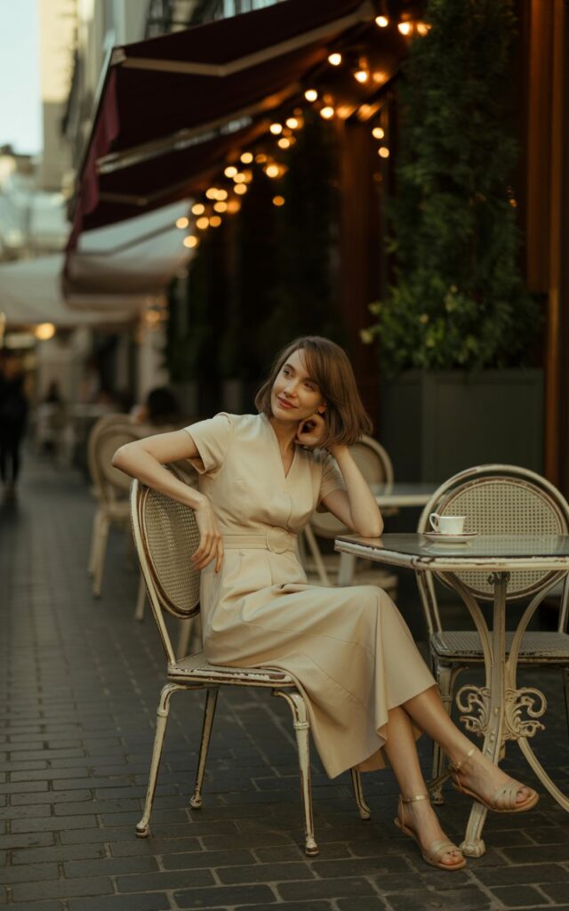 A full-body image at a chic urban café patio with string lights overhead during dusk. The model wears a cream midi dress cinched at the waist with a built-in belt, paired with nude sandals. Her shoulder-length hair is soft and tousled, and she carries no accessories. The soft golden light illuminates her face and dress subtly. She stands relaxed, one arm resting on a chair, with a gentle smile and playful eyes.