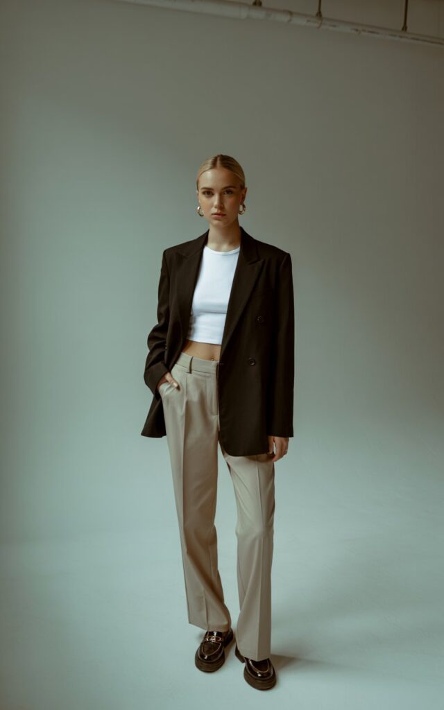 A studio-style fashion photo with soft indoor light. Model stands tall, arms crossed confidently. She wears a sleek black blazer over a white crop top and tailored beige trousers with loafers. Hair in a sleek low bun, gold hoops as accessories. Serious, polished expression.