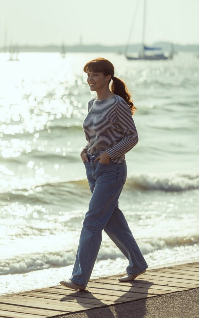 Classic brunette with loose ponytail, wearing straight-leg blue jeans, striped navy-and-white long-sleeve top, and flats. Shot on a boardwalk by the water, soft daylight. She’s strolling with hands in pockets, relaxed smile.