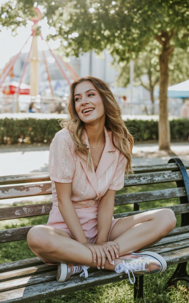 Daytime park setting with carnival rides in the background. White-skinned model wears a pastel pink sparkly romper paired with glitter sneakers. She sits casually on a bench, laughing candidly with one leg tucked under her. Loose waves and soft peach makeup keep it sweet and fun. Light, playful festival vibes.