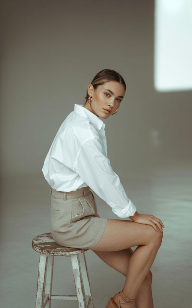 Editorial-style shot in a minimal indoor studio with soft window light. Model wears a crisp oversized white button-up tucked into beige linen shorts with tan strappy sandals. Hair straightened and tucked behind ears. She leans against a wooden stool, confident but relaxed expression.