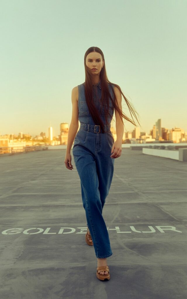 Fit white-skinned model with long, straight dark hair parted down the middle, dressed in a fitted denim jumpsuit with a belted waist, and tan platform sandals. Urban rooftop background at sunset with golden tones. She’s walking forward powerfully, hair flowing naturally, serious expression giving a bold, editorial vibe.