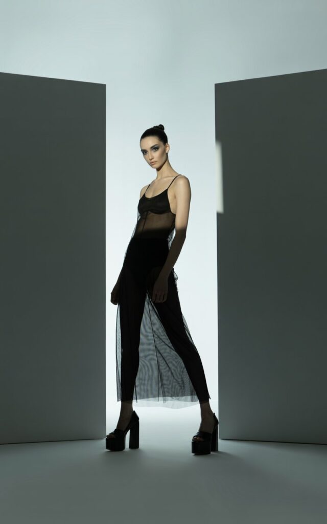 Full-body editorial on a modern minimalist set. Model in a black sheer maxi dress layered over a black bodysuit, paired with platform heels. Hair slick high bun. Makeup smokey eyes. Lighting dramatic side spotlight. Pose standing tall with legs slightly apart, bold gaze.