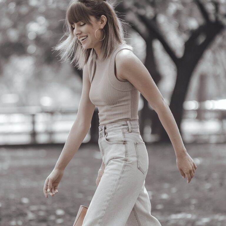 Full-body image in a park setting with leafy trees and soft filtered daylight. The model wears a beige ribbed tank, high-waisted light-wash wide-leg jeans, and tan leather sandals. Large gold hoop earrings accentuate her look, along with a simple tote bag. Her hair is styled in a half-up bun, soft strands falling naturally. She’s captured mid-step, laughing lightly with her head turned toward the camera.