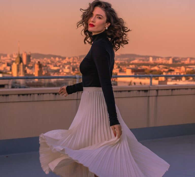 Full-body urban rooftop photo. Model wears a flowy pleated white midi skirt with a slim black long-sleeve top tucked in. Pointed heels and red lipstick finish the look. Hair styled in loose glamorous curls. Golden-hour lighting enhances elegance as she twirls lightly, skirt flowing.