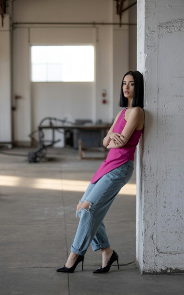 Industrial warehouse backdrop with soft window lighting. Model wears a fitted one-shoulder top in bold color, paired with relaxed ripped boyfriend jeans and heels. Hair straight and sleek. Pose leaning against a wall, arms crossed, edgy and confident.