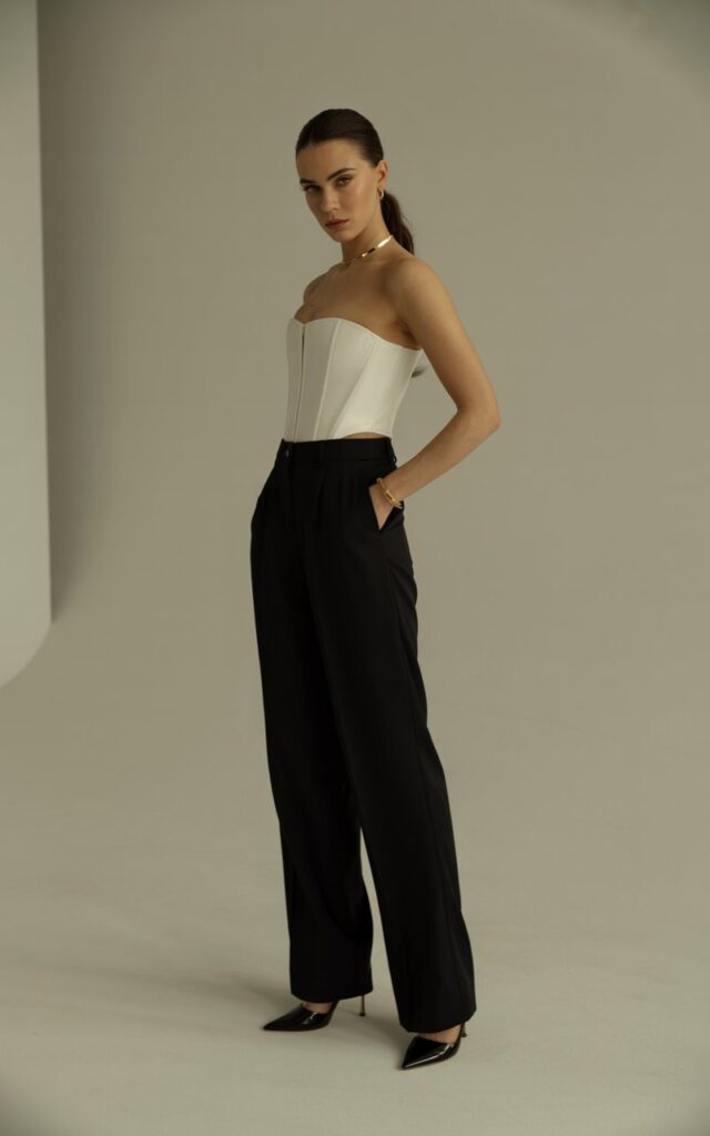 Model wearing a structured white corset top with high-waisted black wide-leg trousers and sleek stilettos. Studio background with soft indoor lighting and a neutral beige wall. Hair pulled back into a sleek ponytail, minimal gold jewelry. Pose standing tall, one hand in pocket, sharp and powerful expression.