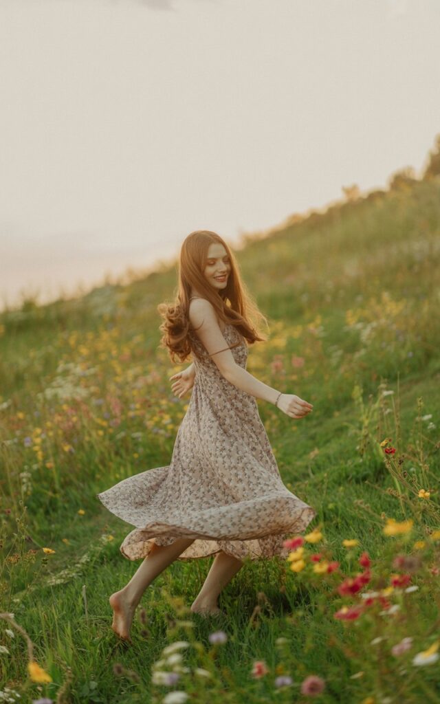Nature setting with wildflowers during golden hour. Model wears a flowing maxi dress in muted floral prints with bare feet in the grass. Hair in soft waves. Warm light catches the fabric as she spins playfully, carefree and romantic.