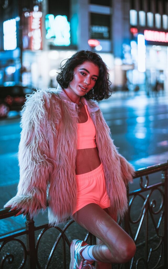 Night street shot with neon signs glowing in the background. Model wears neon pink matching crop top and shorts set layered under a shaggy white faux fur jacket, paired with metallic sneakers. She leans casually on a railing, hair in tousled curls, subtle glossy lips. A mix of luxe and edgy rave style.