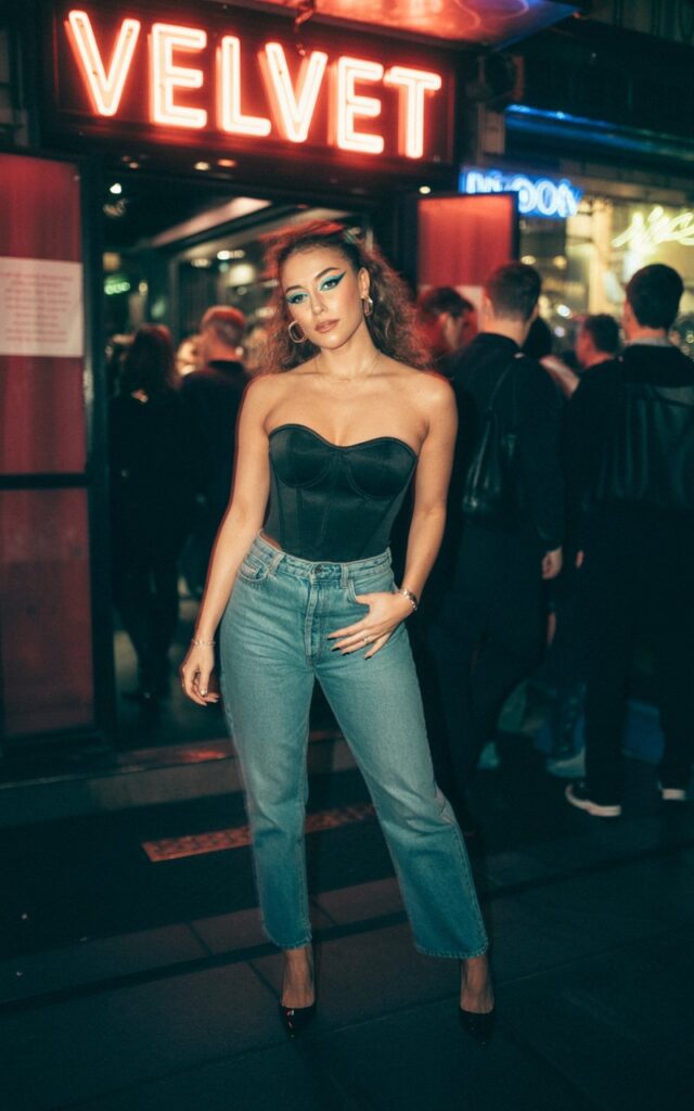 Nighttime club entrance with moody lighting. She wears a structured black corset top with medium-wash straight-leg jeans and pointed heels. Hair in glamorous loose curls, makeup bold with winged eyeliner. She’s standing confidently with one hand on her hip.