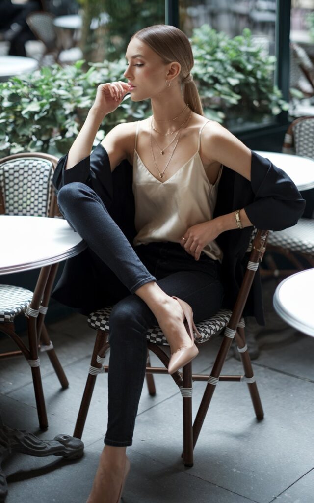 Outdoor café scene. Model in dark skinny jeans with a champagne silk cami, nude heels, and layered dainty necklaces. Hair in a sleek ponytail, minimal makeup. Natural window light on her face as she sits at a bistro chair, crossing her legs elegantly.