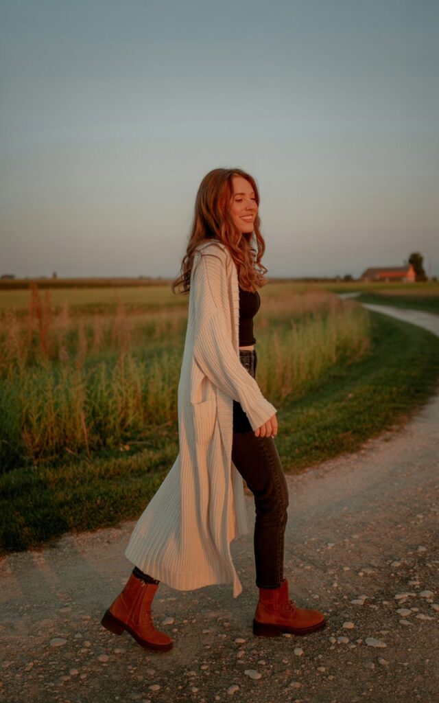 Rustic countryside path. Model in a floor-length knit duster cardigan over a fitted top and jeans, paired with suede booties. Golden hour sunset glow. Hair flowing freely in the breeze. Pose walking away but glancing back with a warm smile.