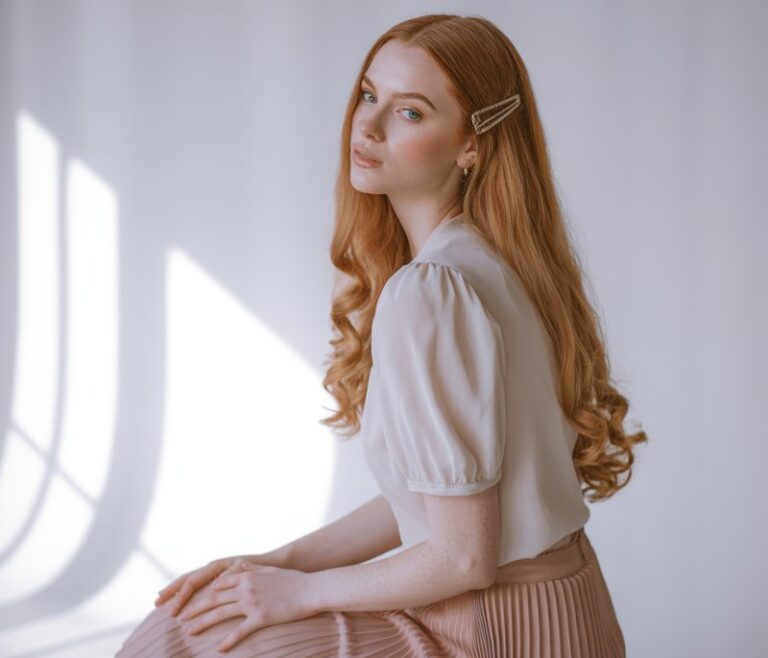Slender fair-skinned model with long auburn hair in soft curls, wearing a blush pink pleated midi skirt, tucked white puff-sleeve blouse, and nude heels. Studio background with soft window light streaming in. She’s seated gracefully on a stool, hands resting on her lap, dreamy expression.